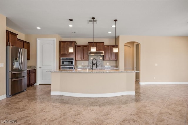 kitchen with tasteful backsplash, light stone counters, an island with sink, decorative light fixtures, and appliances with stainless steel finishes