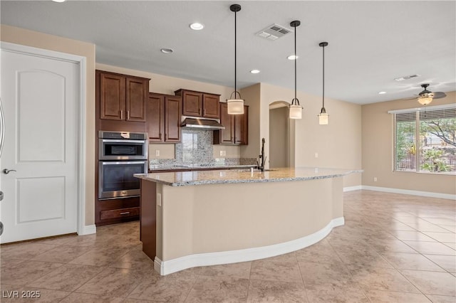 kitchen featuring light stone countertops, backsplash, double oven, a kitchen island with sink, and pendant lighting