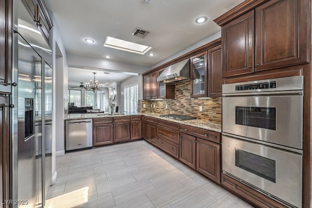 kitchen with a skylight, wall chimney range hood, an inviting chandelier, pendant lighting, and appliances with stainless steel finishes