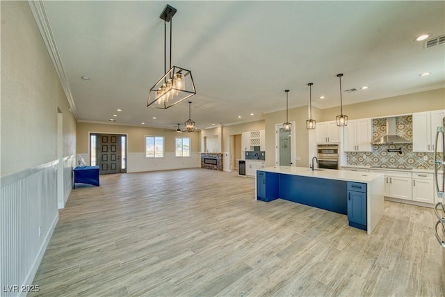 kitchen featuring pendant lighting, white cabinetry, blue cabinetry, a center island with sink, and wall chimney exhaust hood