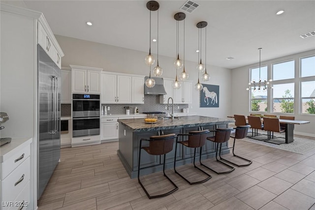 kitchen featuring a kitchen bar, appliances with stainless steel finishes, a kitchen island with sink, wall chimney range hood, and white cabinetry