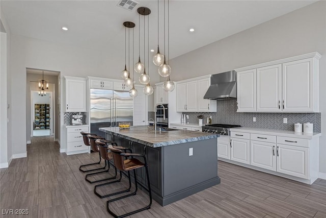 kitchen featuring a kitchen breakfast bar, wall chimney exhaust hood, a kitchen island with sink, white cabinetry, and hanging light fixtures