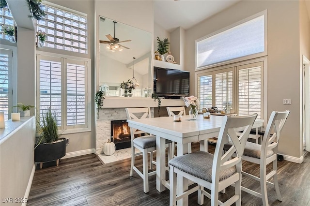 dining room with a stone fireplace, ceiling fan, high vaulted ceiling, and dark wood-type flooring