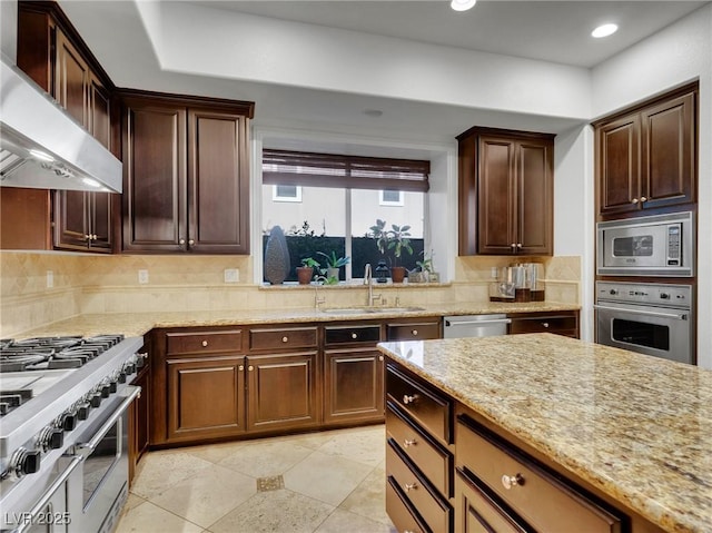 kitchen featuring light stone counters, wall chimney range hood, backsplash, appliances with stainless steel finishes, and sink