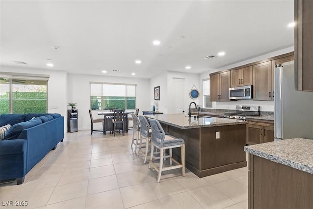 kitchen with a kitchen island with sink, a breakfast bar area, light tile patterned floors, stone countertops, and stainless steel appliances