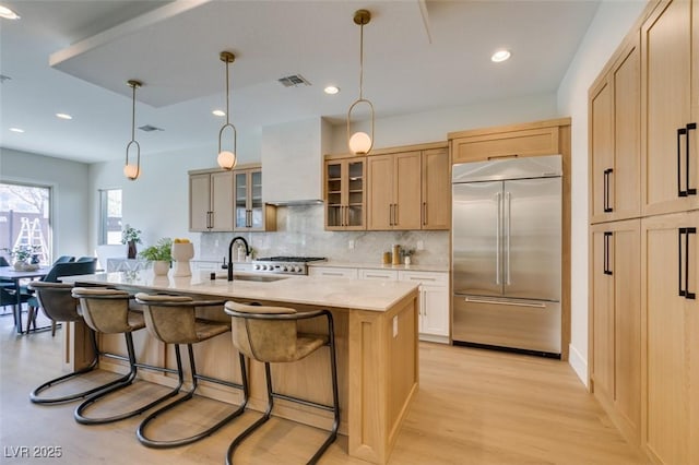 kitchen featuring a center island with sink, sink, built in refrigerator, light brown cabinetry, and custom range hood