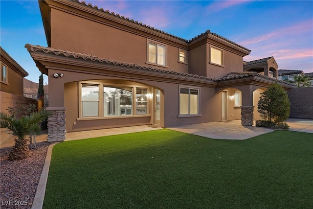 back of house at dusk with a lawn, stone siding, a tile roof, a patio area, and stucco siding