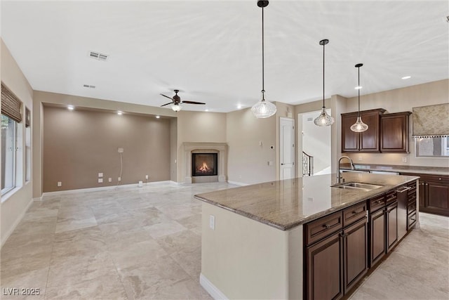 kitchen with decorative light fixtures, a sink, an island with sink, light stone countertops, and a warm lit fireplace