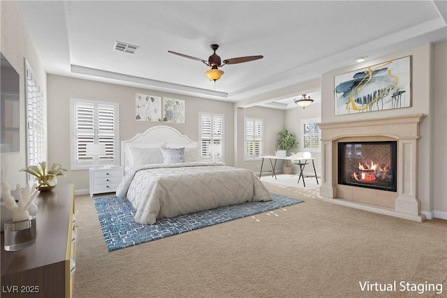 carpeted bedroom with ceiling fan, visible vents, baseboards, a tray ceiling, and a glass covered fireplace