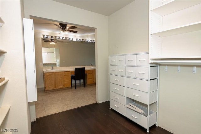 spacious closet with built in desk, sink, ceiling fan, and dark wood-type flooring