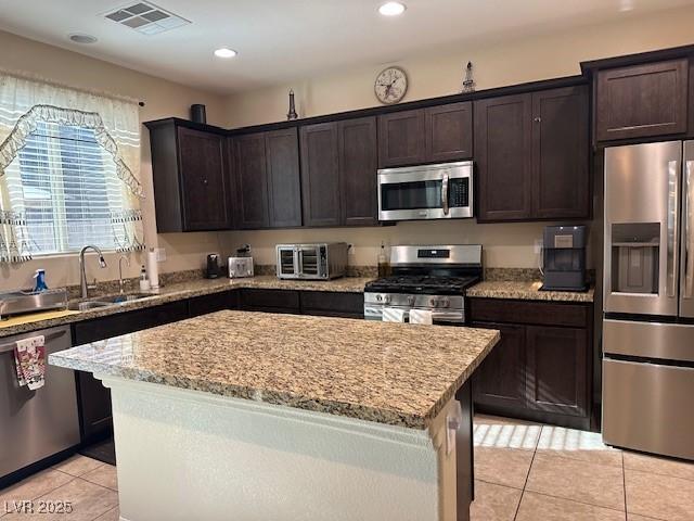 kitchen featuring dark brown cabinetry, a center island, light tile patterned floors, stainless steel appliances, and light stone countertops