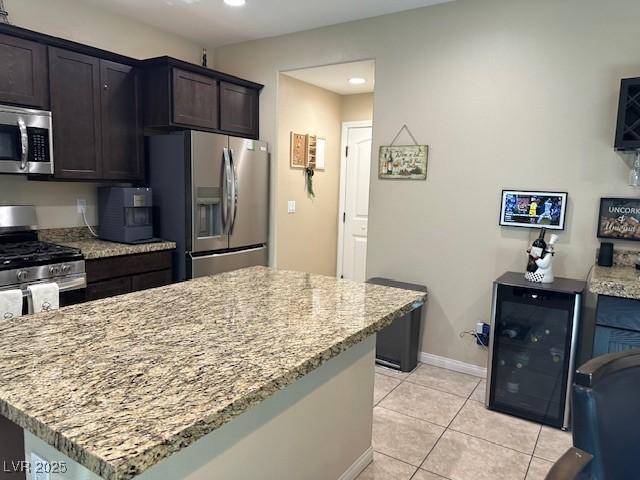 kitchen with appliances with stainless steel finishes, beverage cooler, light tile patterned floors, dark brown cabinetry, and light stone counters