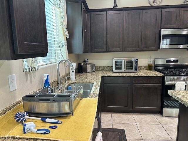 kitchen with sink, light tile patterned floors, appliances with stainless steel finishes, dark brown cabinetry, and light stone counters