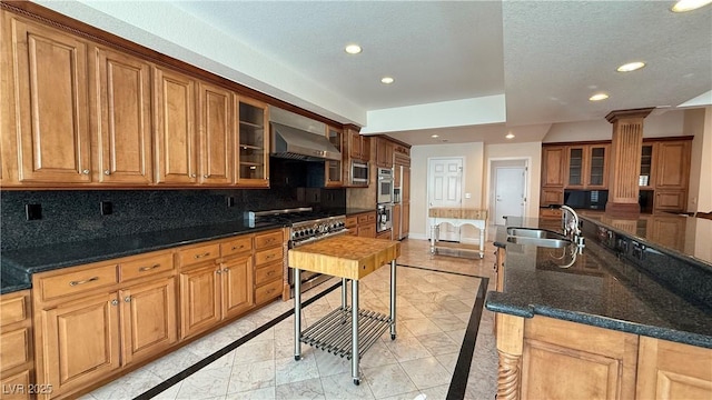 kitchen featuring sink, wall chimney exhaust hood, decorative columns, dark stone counters, and appliances with stainless steel finishes