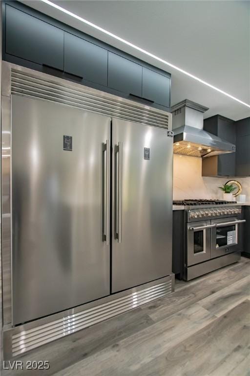 kitchen featuring wall chimney exhaust hood, high quality appliances, tasteful backsplash, and light hardwood / wood-style flooring