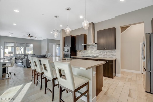 kitchen with appliances with stainless steel finishes, wall chimney exhaust hood, a breakfast bar, dark brown cabinets, and a kitchen island with sink