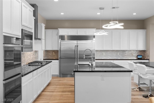 kitchen featuring appliances with stainless steel finishes, light wood-type flooring, decorative light fixtures, white cabinets, and a kitchen island with sink