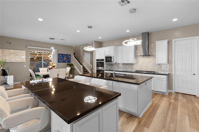 kitchen with wall chimney range hood, an island with sink, white cabinetry, and decorative light fixtures