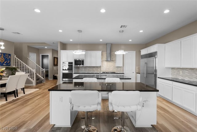 kitchen featuring a kitchen island with sink, stainless steel appliances, wall chimney range hood, and pendant lighting