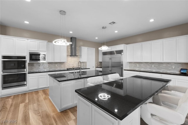 kitchen with a center island with sink, hanging light fixtures, stainless steel appliances, wall chimney range hood, and white cabinetry