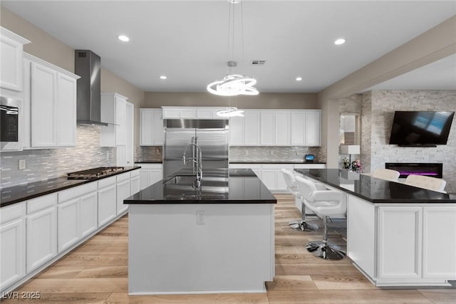 kitchen with a center island with sink, appliances with stainless steel finishes, pendant lighting, white cabinetry, and wall chimney range hood