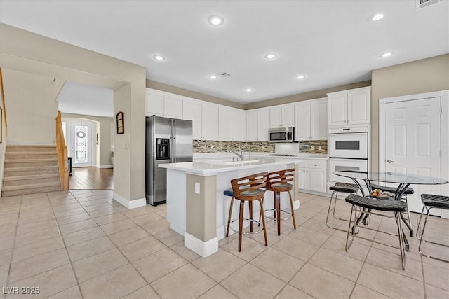 kitchen featuring stainless steel appliances, light tile patterned flooring, backsplash, an island with sink, and white cabinets