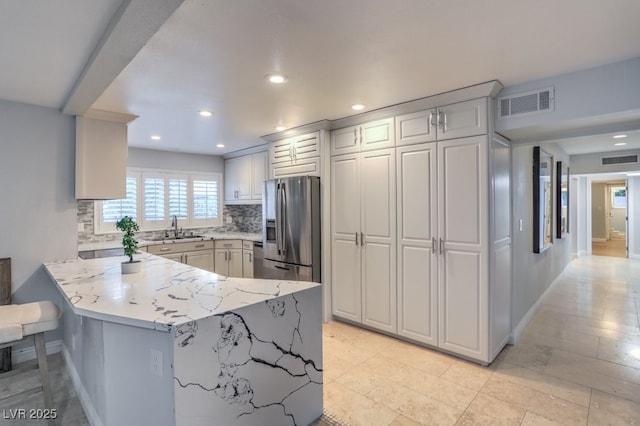 kitchen featuring kitchen peninsula, stainless steel refrigerator with ice dispenser, tasteful backsplash, light stone counters, and sink