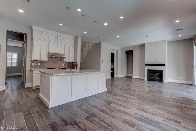 kitchen with white cabinets, under cabinet range hood, visible vents, baseboards, and a kitchen island with sink