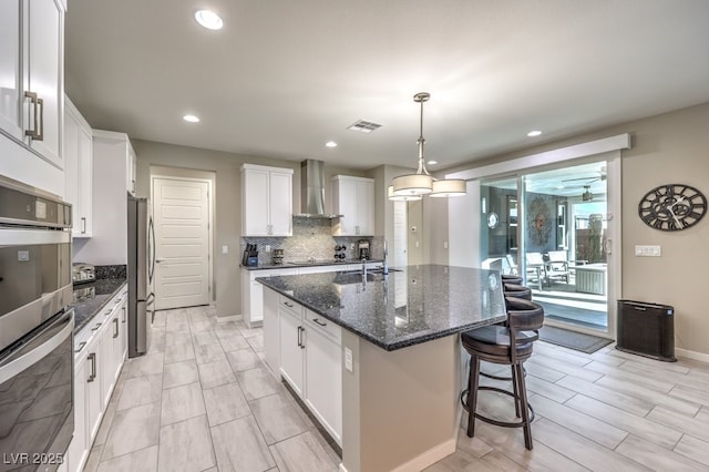 kitchen with wall chimney exhaust hood, stainless steel appliances, pendant lighting, a center island with sink, and white cabinets