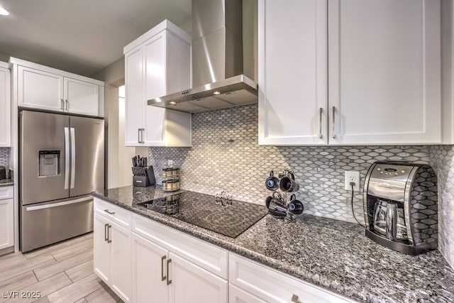 kitchen featuring black electric stovetop, wall chimney range hood, stainless steel fridge, dark stone countertops, and white cabinetry