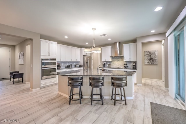 kitchen featuring dark stone counters, white cabinets, sink, wall chimney exhaust hood, and an island with sink