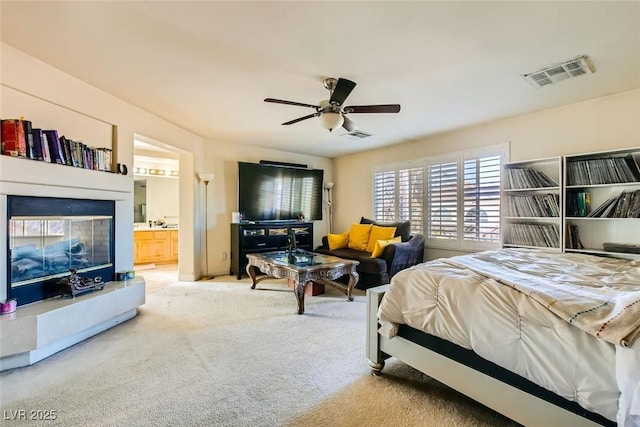 bedroom featuring ceiling fan, ensuite bath, a tiled fireplace, and carpet flooring