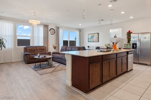 kitchen with dark brown cabinetry, stainless steel appliances, decorative light fixtures, a center island with sink, and a notable chandelier