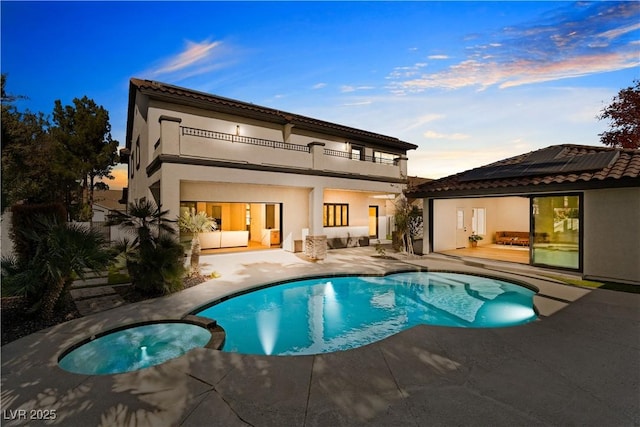 back house at dusk with solar panels, a balcony, a patio, and a pool with hot tub