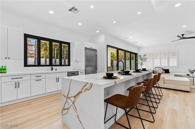 kitchen with stainless steel built in refrigerator, white cabinets, light stone countertops, and an island with sink