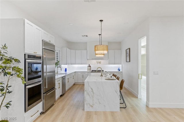 kitchen featuring appliances with stainless steel finishes, light wood-type flooring, white cabinetry, hanging light fixtures, and an island with sink