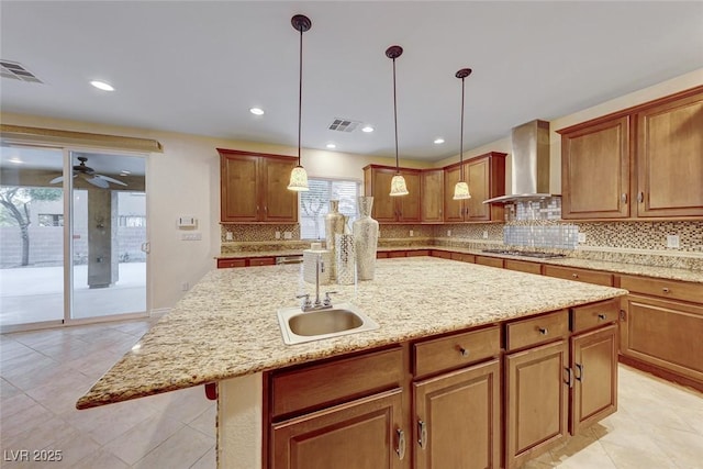 kitchen with wall chimney exhaust hood, an island with sink, stainless steel gas stovetop, ceiling fan, and sink