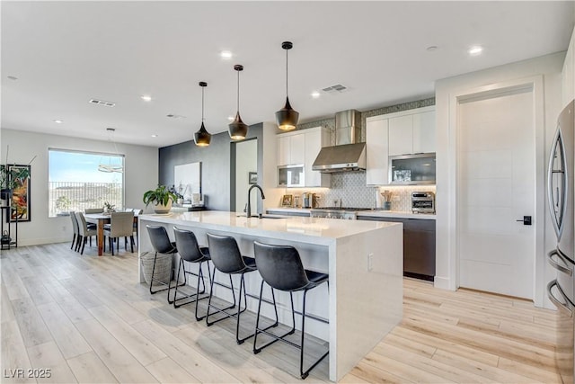 kitchen with backsplash, a large island with sink, wall chimney range hood, hanging light fixtures, and white cabinetry