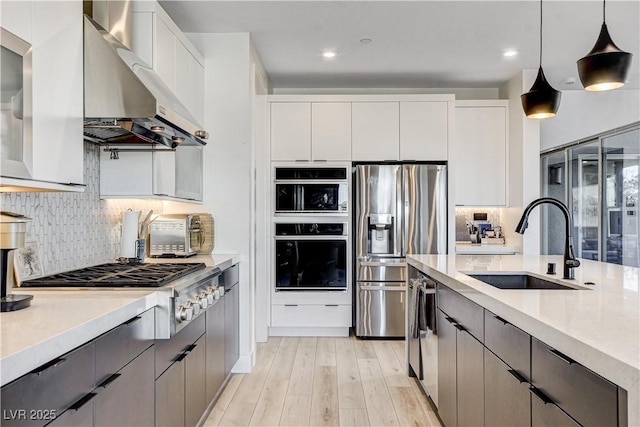 kitchen featuring wall chimney exhaust hood, stainless steel appliances, sink, white cabinetry, and hanging light fixtures