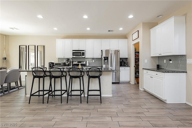 kitchen with a kitchen bar, stainless steel appliances, dark stone countertops, white cabinetry, and an island with sink