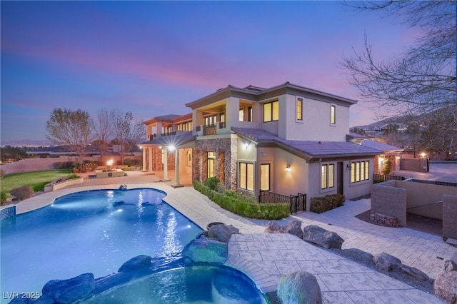 back house at dusk with pool water feature, a patio area, a swimming pool with hot tub, and a balcony