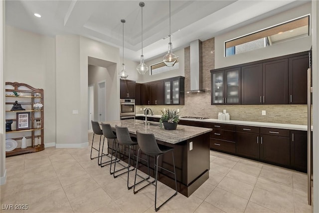 kitchen featuring wall chimney range hood, a raised ceiling, a towering ceiling, an island with sink, and gas stovetop