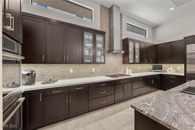 kitchen featuring dark brown cabinetry, appliances with stainless steel finishes, wall chimney exhaust hood, backsplash, and light tile patterned floors