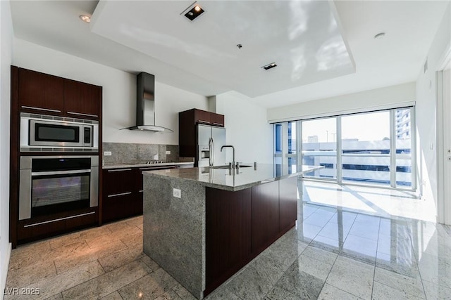 kitchen with backsplash, stone counters, a kitchen island with sink, wall chimney exhaust hood, and appliances with stainless steel finishes