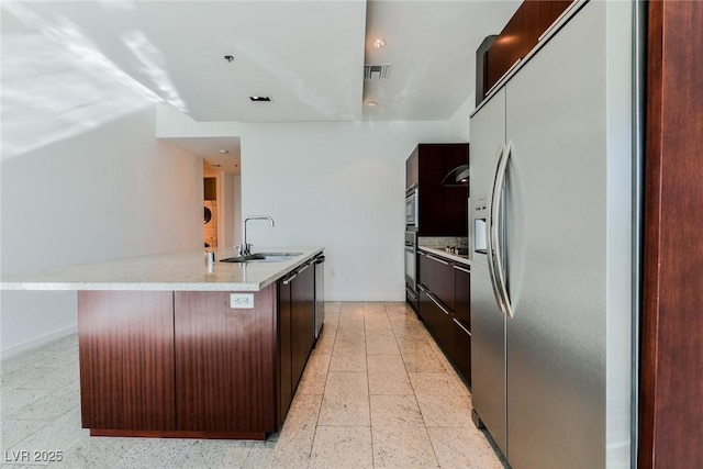 kitchen with sink, an island with sink, appliances with stainless steel finishes, light stone counters, and dark brown cabinetry