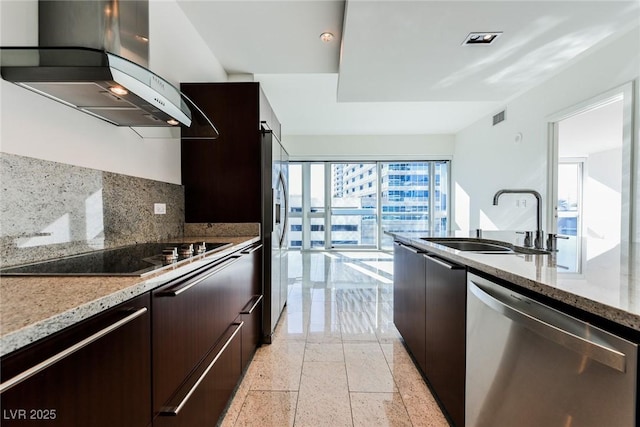 kitchen featuring backsplash, light stone counters, wall chimney exhaust hood, stainless steel appliances, and sink