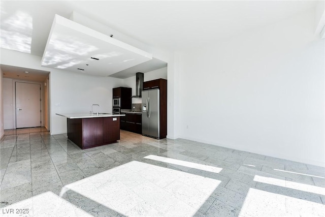 kitchen featuring appliances with stainless steel finishes, dark brown cabinets, a kitchen island with sink, wall chimney range hood, and sink