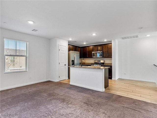 kitchen featuring light stone counters, light colored carpet, appliances with stainless steel finishes, and a kitchen island with sink