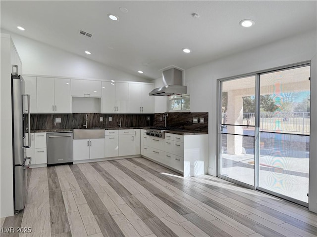 kitchen featuring wall chimney range hood, sink, white cabinetry, stainless steel appliances, and vaulted ceiling