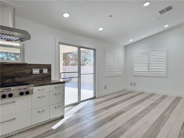 kitchen featuring wall chimney range hood, backsplash, white cabinets, stainless steel gas stovetop, and light wood-type flooring